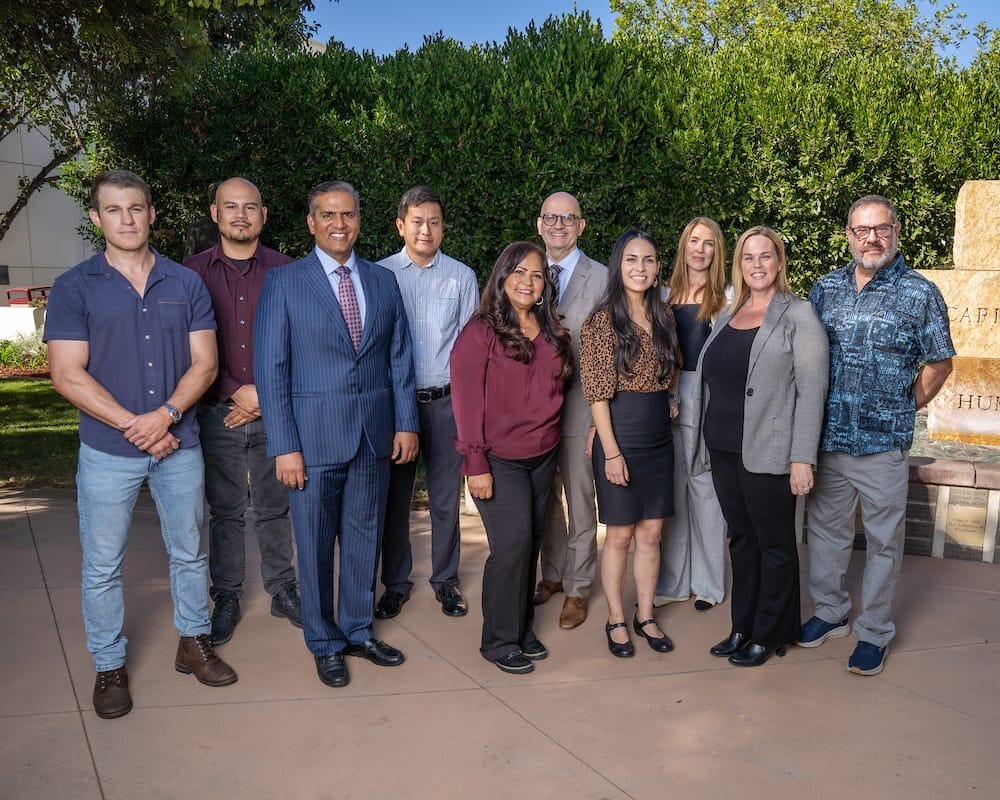 A group of ten people standing outdoors in front of a stone sign with trees in the background.